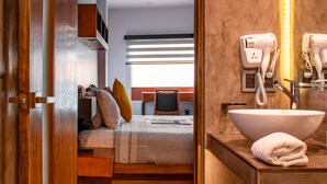 A hotel room with a wooden door, a bed with pillows, a bedside table, a bathroom with a sink, a mirror, and a hairdryer. - Madero Hotel & Suites (La Paz)