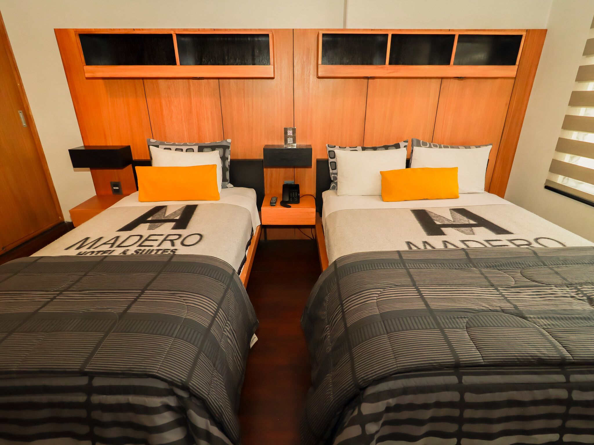 Habitación de hotel con dos camas, cada una con colcha de marca 'MADERO', cabeceros de madera y una mesita de noche con lámpara.