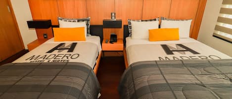 Habitación de hotel con dos camas, cada una con colcha de marca 'MADERO', cabeceros de madera y una mesita de noche con lámpara.