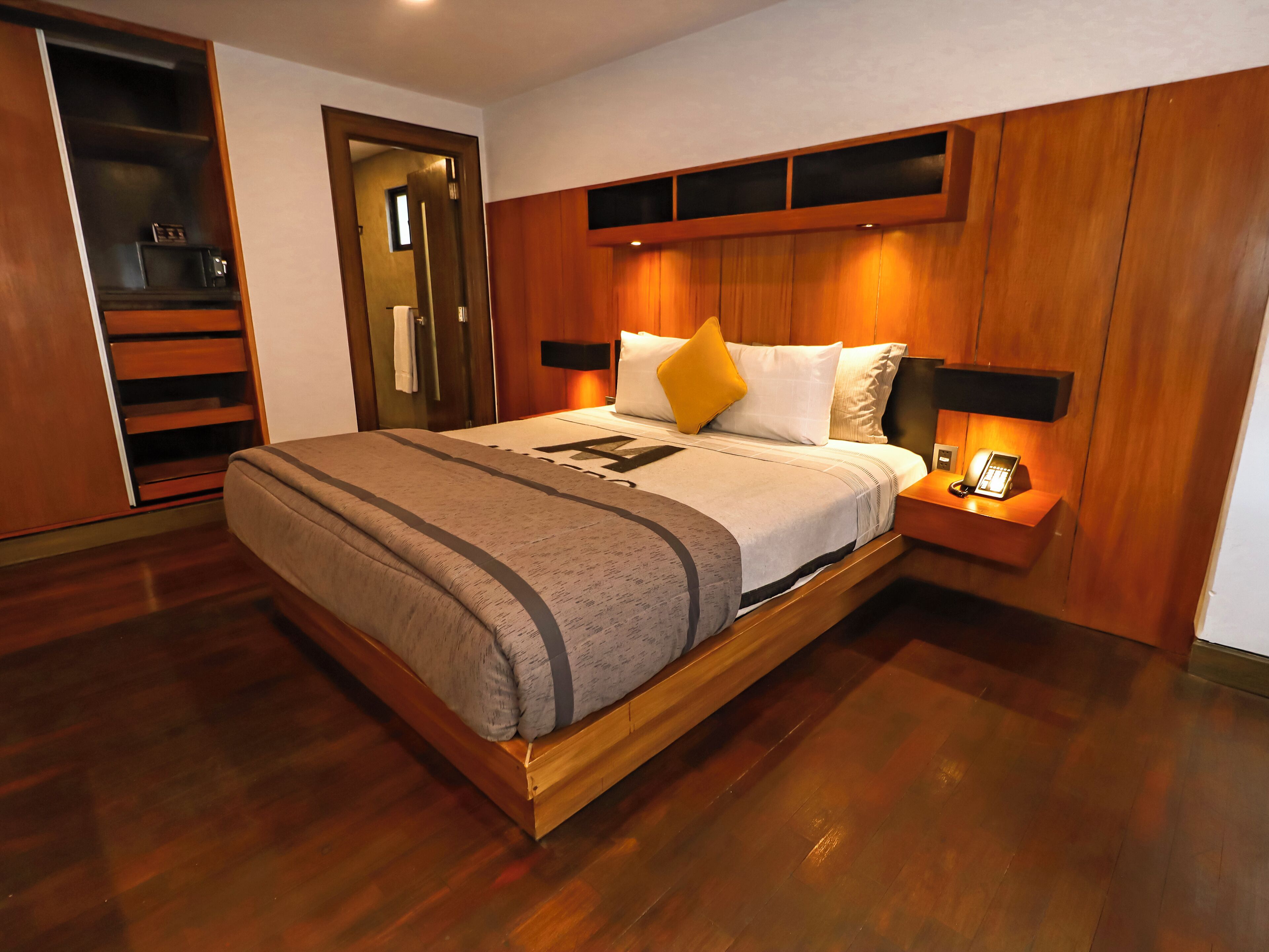 A modern hotel room with a wooden bed frame, a built-in wardrobe, and a bedside table with a lamp.