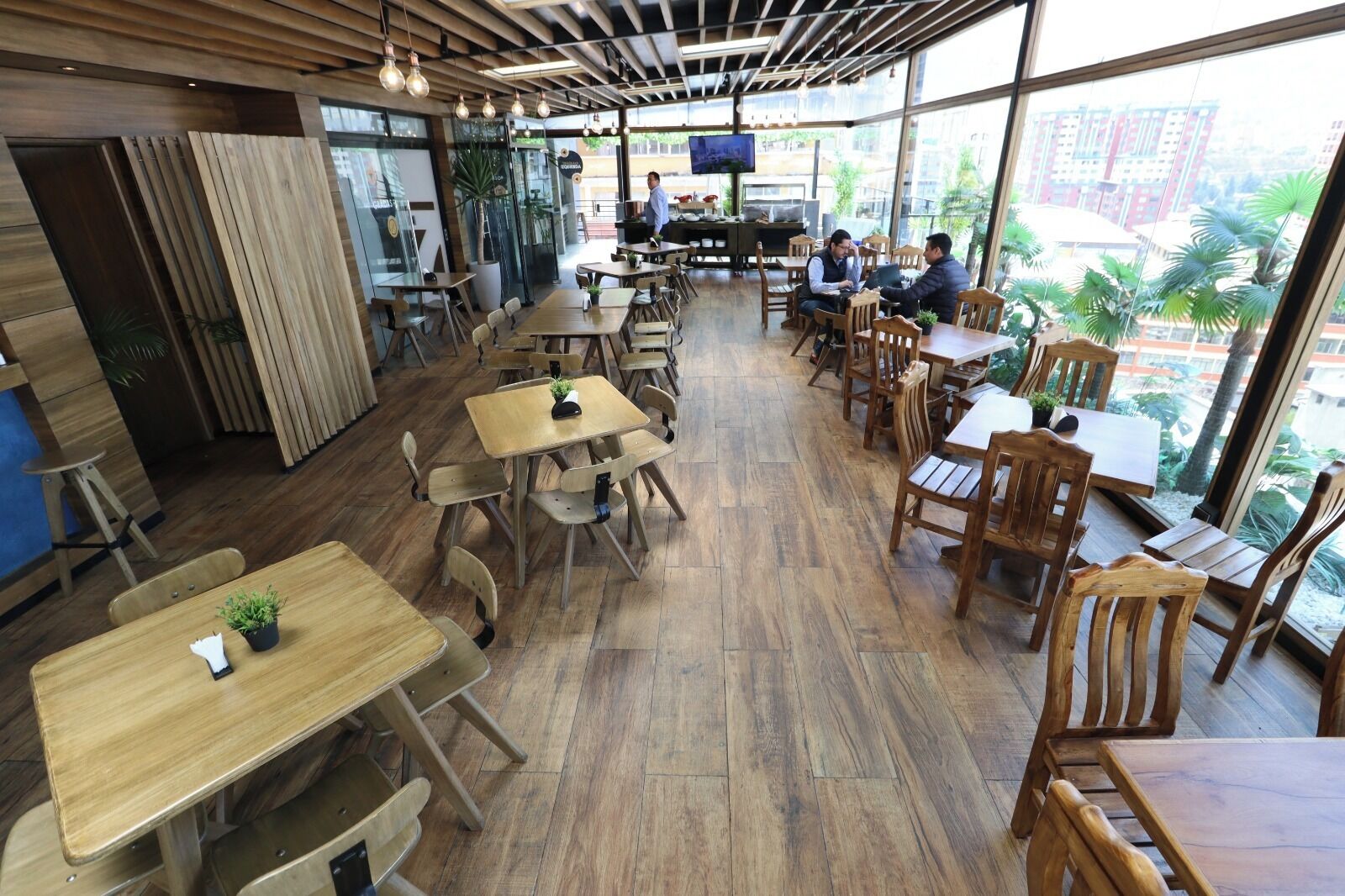 A spacious cafe with wooden tables and chairs, large windows offering a view of the city, and a modern interior design.