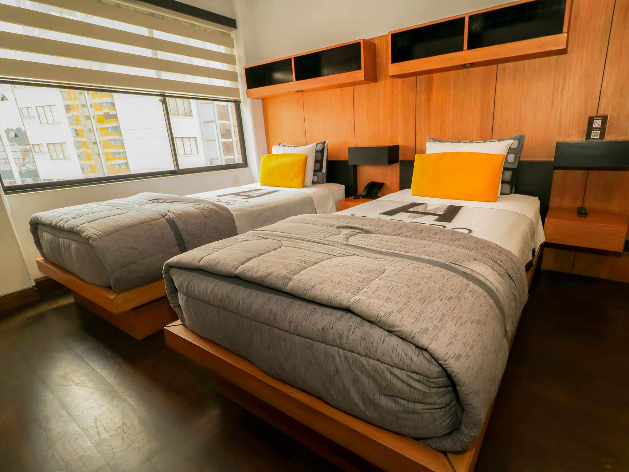 A modern hotel room with two beds, wooden headboards, and a large window with blinds.
