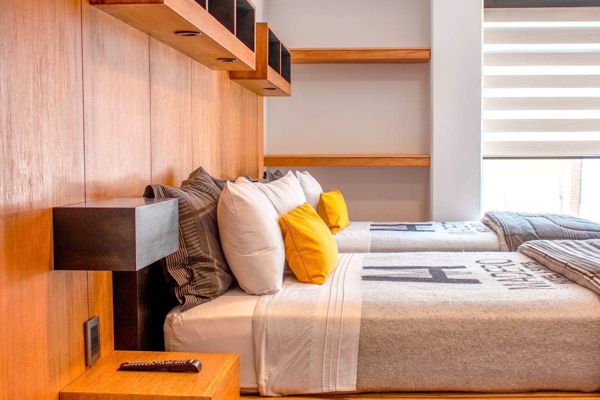 A modern bedroom with a wooden headboard, a bedside table with a remote control, and a window with blinds.