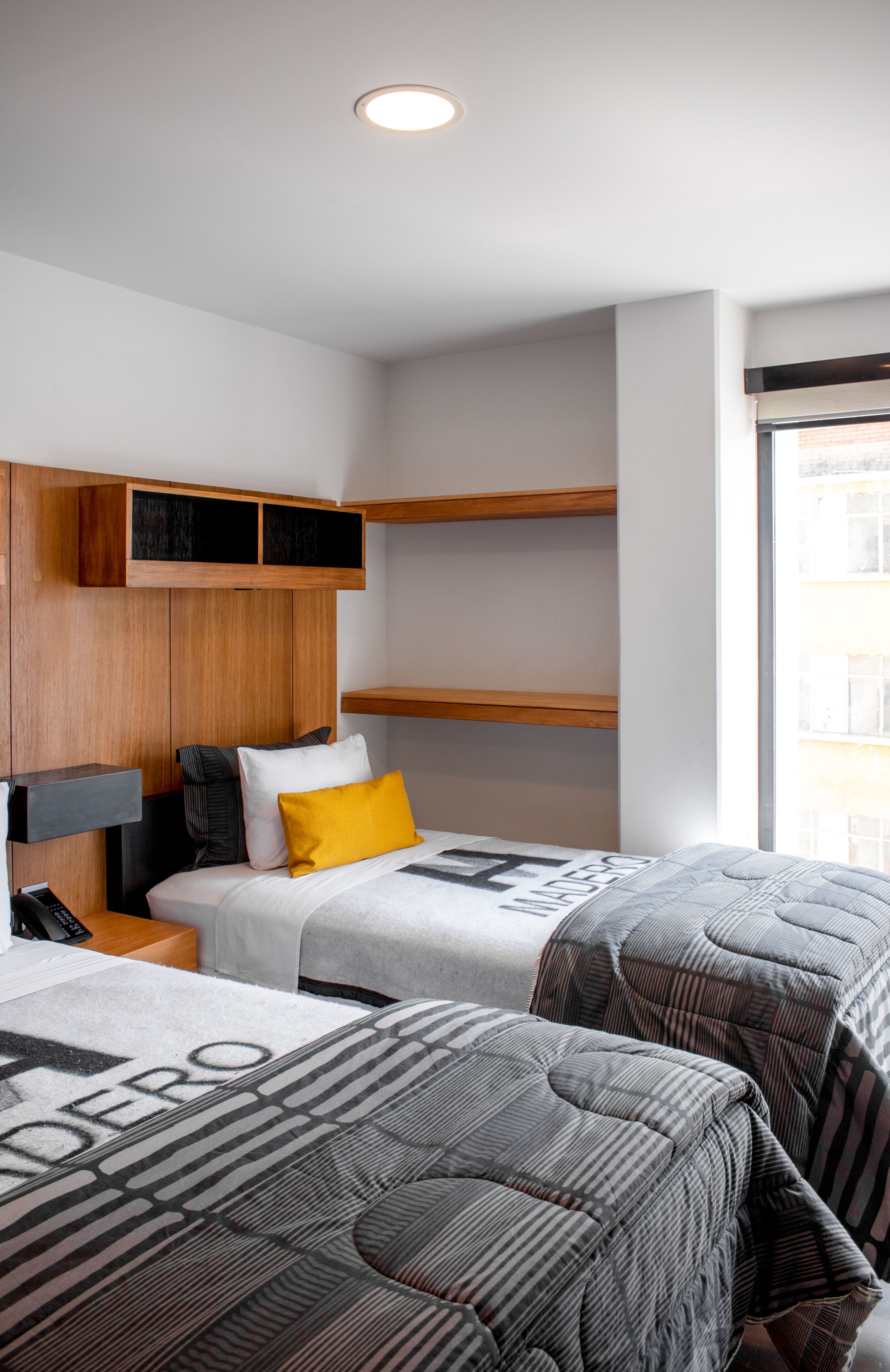 A hotel room with two beds, wooden headboards, a bedside table with a lamp, and a window with a view of buildings.