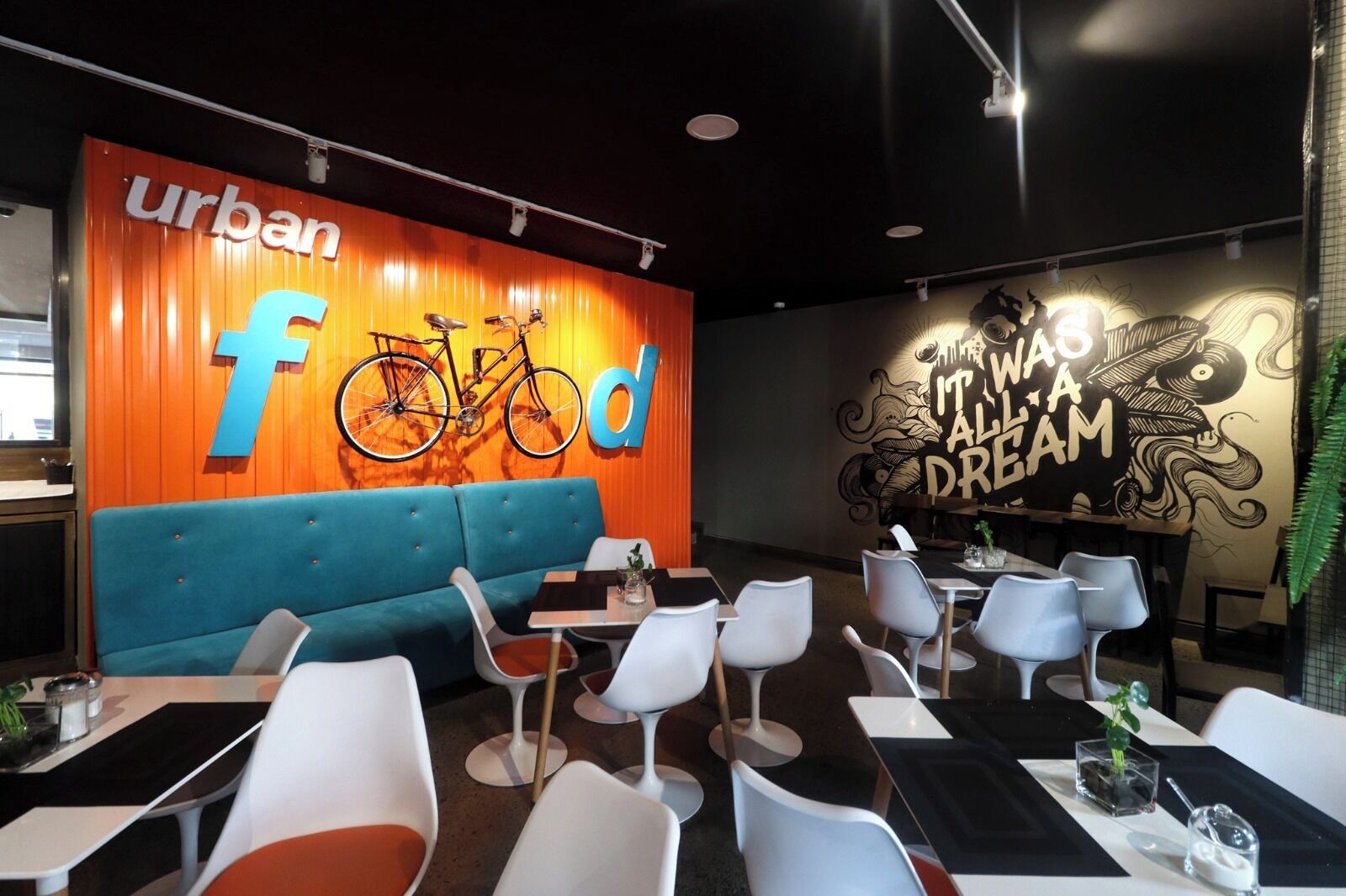 Ein Restaurantinnenraum mit einem blauen Sofa, weißen Stühlen und Tischen. Ein großes Wandbild zeigt den Schriftzug 'urban food' und ein Fahrrad.