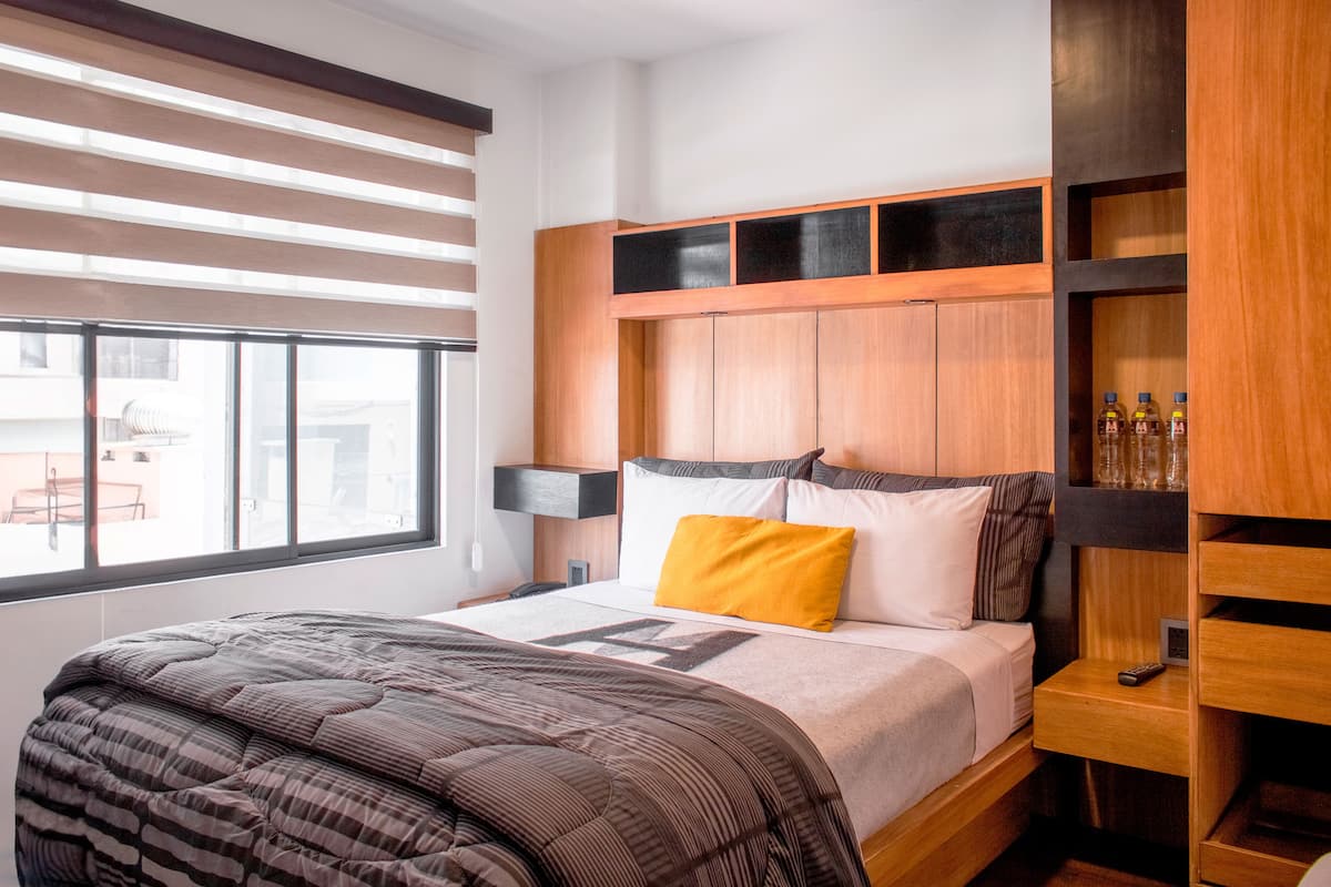 A modern hotel room with a large bed, a nightstand, a wooden headboard, and a window with blinds.