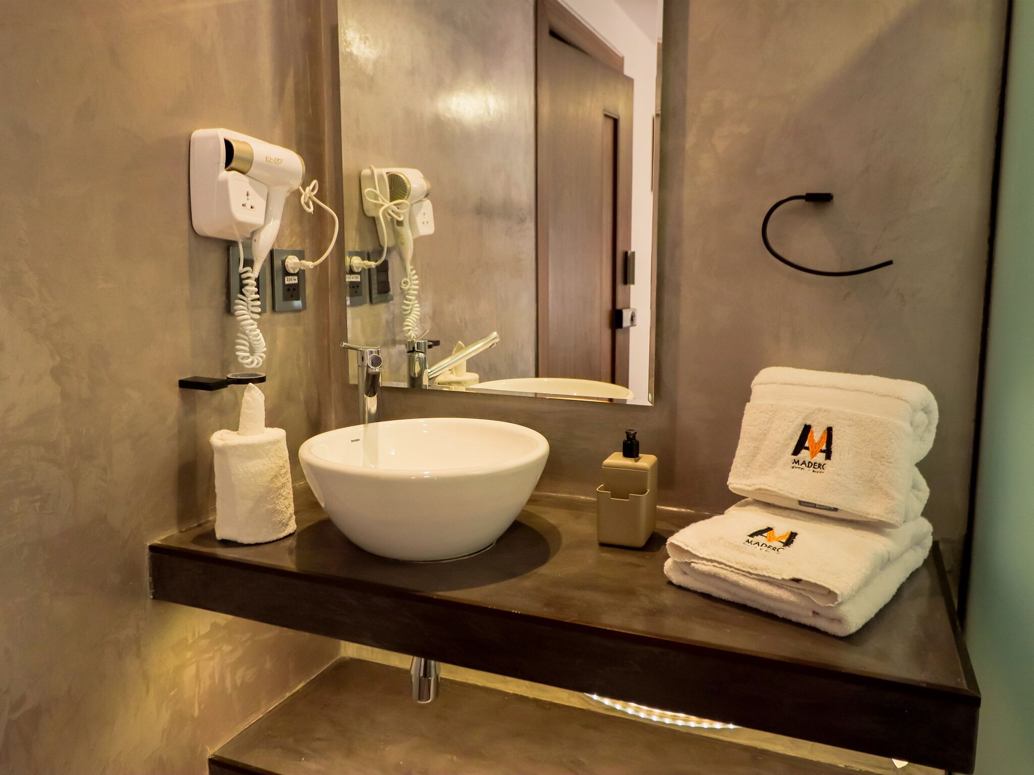 Ein modernes Badezimmer mit einem großen weißen Waschbecken, einem Spiegel und zwei an der Wand montierten Haartrocknern. Auf der Ablage befinden sich ein Handtuchspender und eine Flasche Seife.