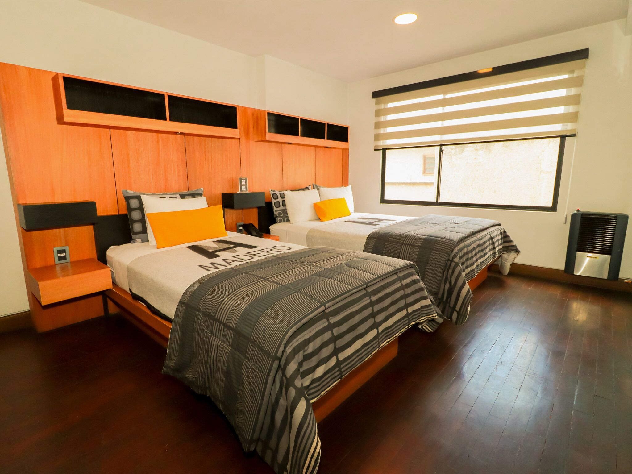 A hotel room with two beds, wooden headboards, a large window with blinds, and a built-in air conditioning unit.