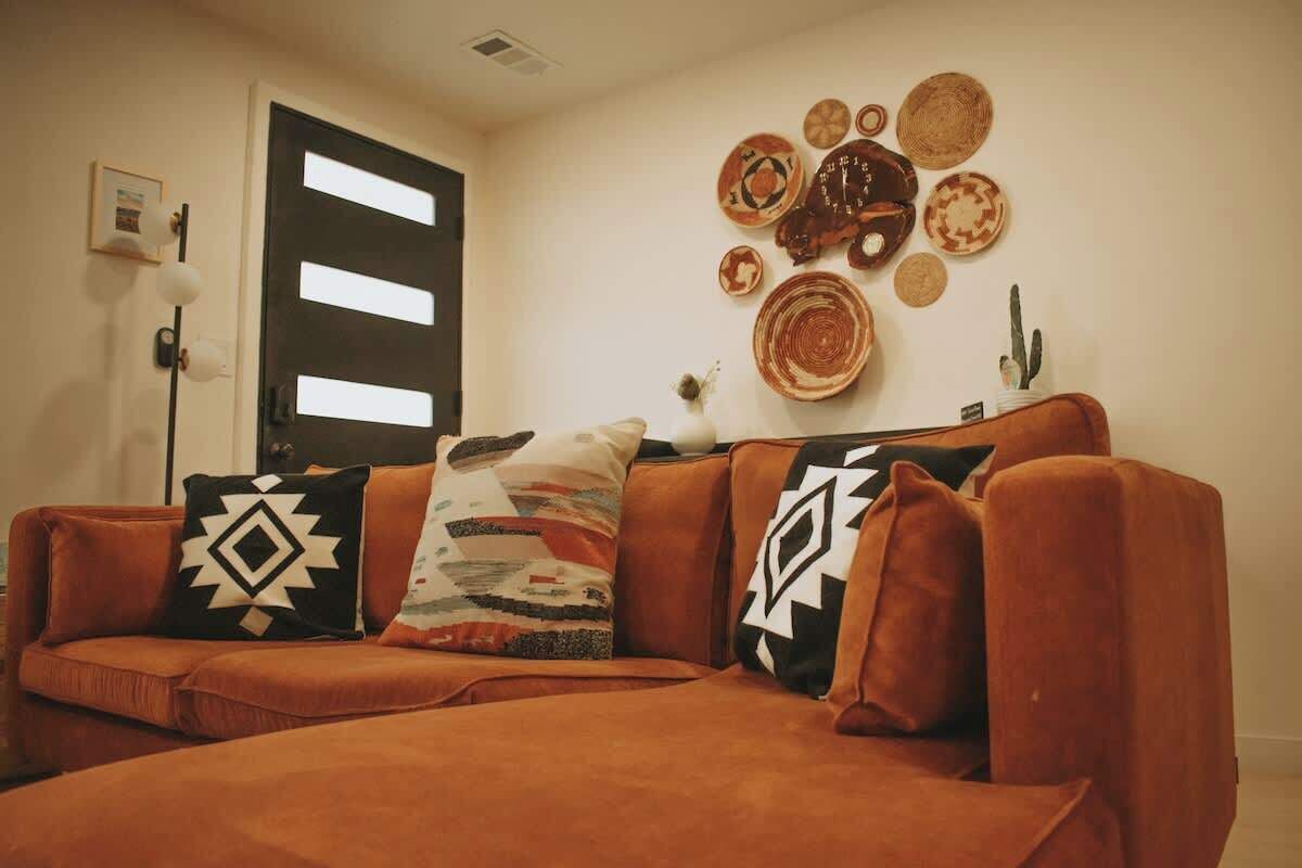 A living room with a brown sofa, patterned cushions, and a wall decorated with woven plates and a cactus.
