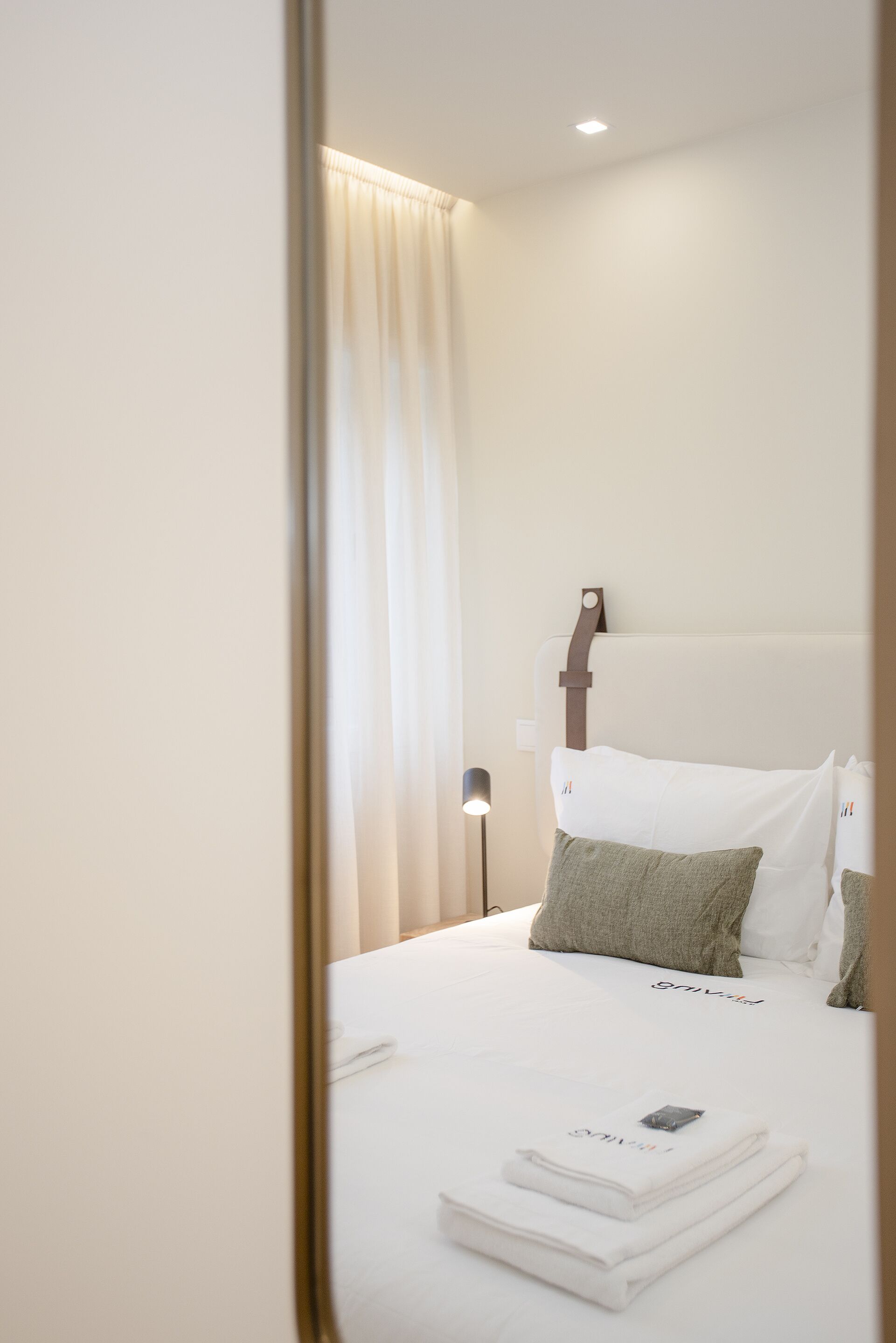 A neatly made bed with white linens, a headboard with a brown strap, and a bedside lamp.