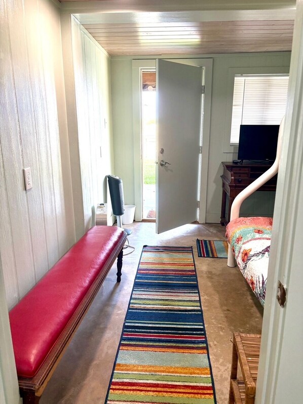 A room with a red bench, a colorful striped rug, a bed with a floral bedspread, a television, and a wooden desk.