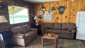 A cabin interior with two brown sofas, a wooden coffee table, a lamp, a sign reading 'Welcome Cabin,' and a 'No Smoking' sign. - Waterfront Cabin with lighted fishing pier (Matagorda)