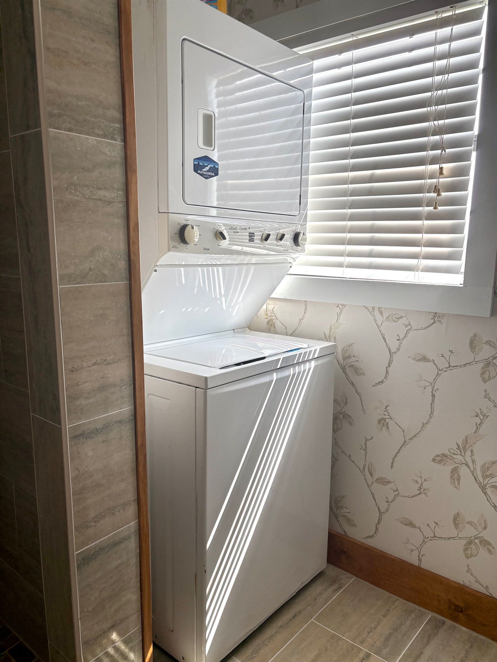 A compact laundry room with a front-loading washer and dryer, a window with blinds, and a tiled backsplash.