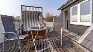 A deck with two chairs and a table, a striped beach umbrella, and a window with blinds. - Ferienwohnung Dünenzauber (St.Peter Ording)
