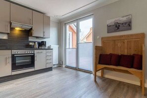 A modern kitchen with an oven, a coffee maker, and a kettle. There is a wooden bench with cushions and a window with blinds.