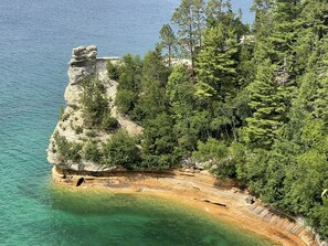 A coastal landscape with a rocky cliff, clear turquoise waters, and dense green trees. - Private hot tub -2 king beds -near Pictured Rocks! (Christmas)