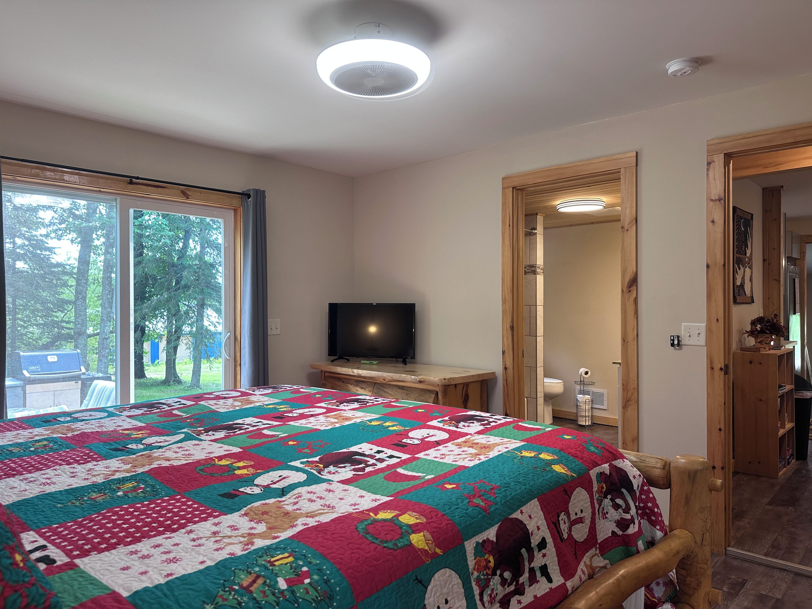 A bedroom with a large bed, a television, a wooden desk, and a sliding glass door leading to a balcony.