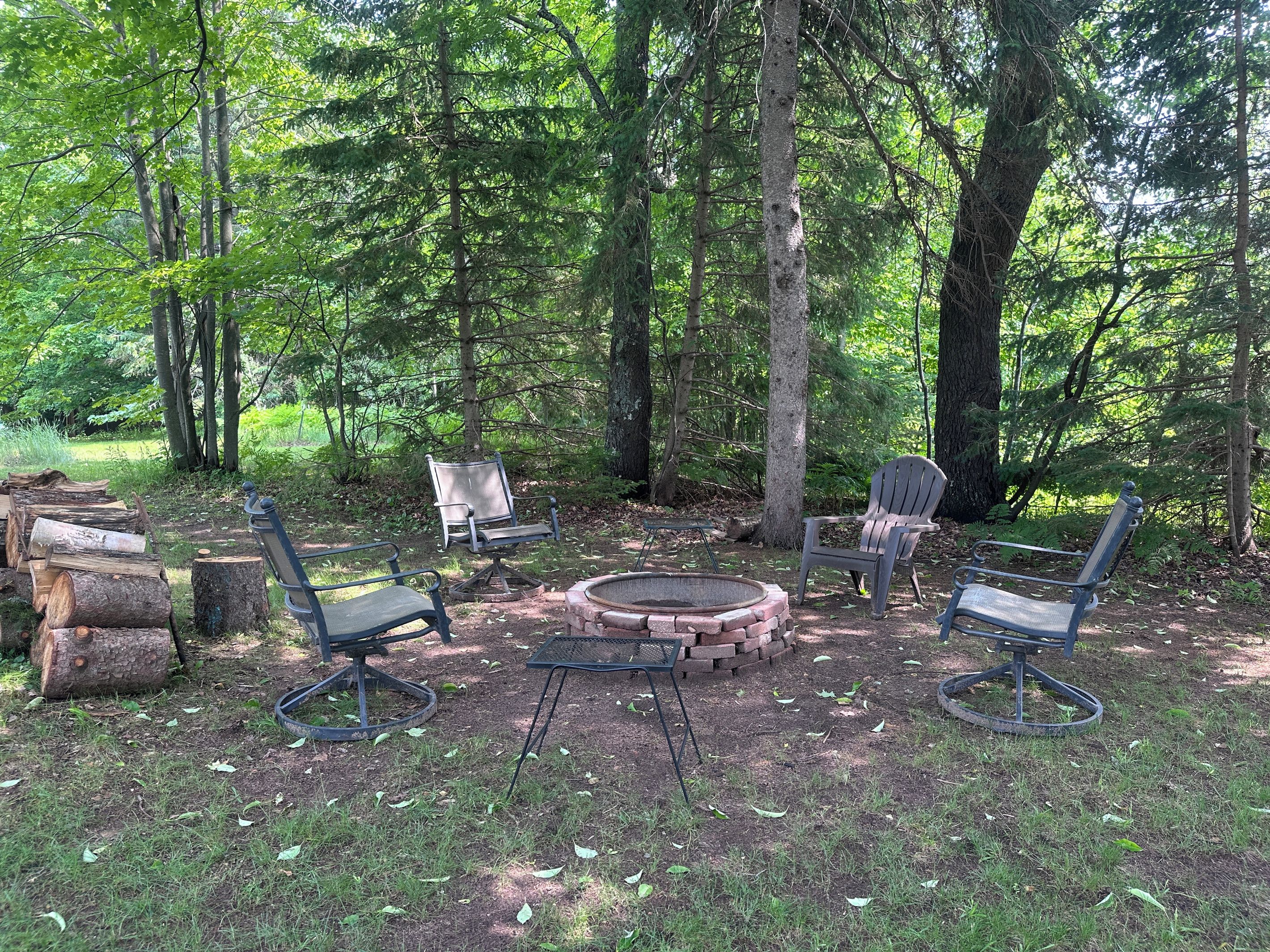 A forested campsite with a fire pit, four Adirondack chairs, and a metal table.