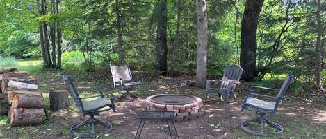 A forested campsite with a fire pit, four Adirondack chairs, and a metal table.