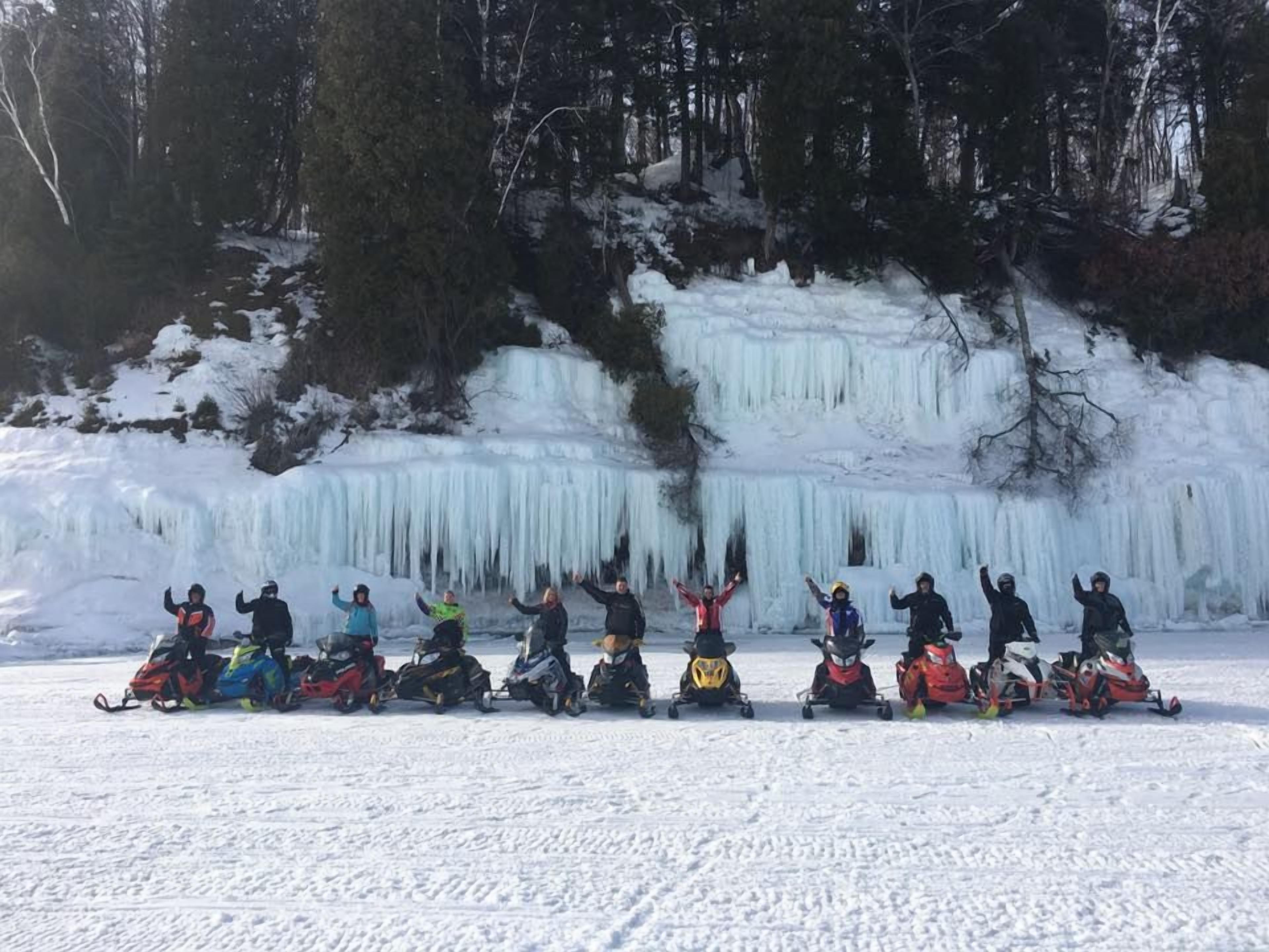 A group of people on snowmobiles in a snowy landscape with a frozen waterfall in the background.