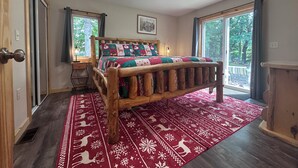 Een houten bed met een sprei, een rood, patroonrijk vloerkleed en uitzicht op groen.