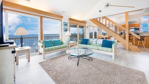 A spacious living room with a large window overlooking the sea, a glass coffee table, and a staircase leading to an upper level.