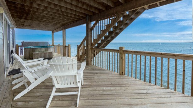 A wooden deck with white Adirondack chairs, a hot tub, and a staircase leading to a balcony overlooking the ocean.