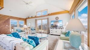 A spacious living room with a high ceiling, large windows, and a view of the sea. There is a ceiling fan, a TV, and a dining area with a glass table.