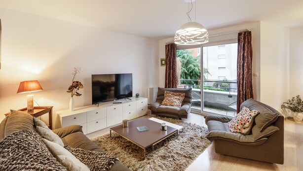 A modern living room with a sofa, coffee table, TV, and a large window with curtains. - Le Bonnet Rouge - Appartement 2 chambres avec parking (Rennes)