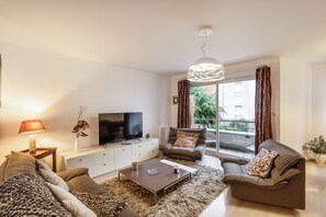 A modern living room with a sofa, coffee table, TV, and a large window with curtains. - Le Bonnet Rouge - Appartement 2 chambres avec parking (Rennes)