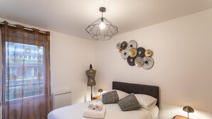 A neatly made bed with pillows and a folded towel, a geometric pendant light, a wall art piece, and a bedside lamp. - Le Bonnet Rouge - Appartement 2 chambres avec parking (Rennes)