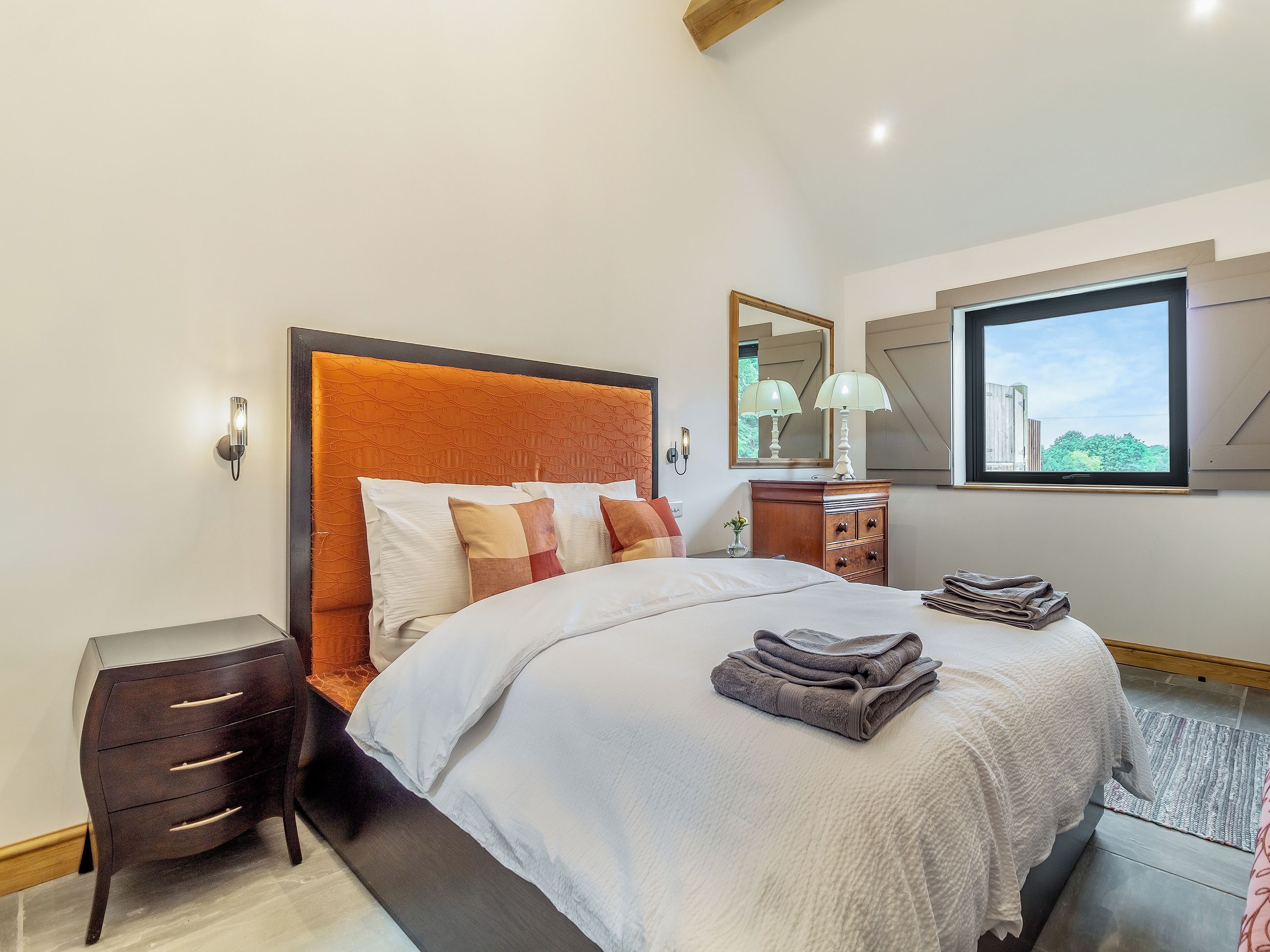 Une chambre moderne avec un grand lit, une table de chevet, une commode et une fenêtre donnant sur un paysage verdoyant.