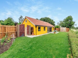 Een geel huis met een rode pannendak, een omheind gazon en een houten terras.