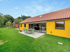 Een geel gebouw met een pannendak, een terras met tuinmeubels en een verzorgd gazon.