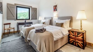 A hotel room with two beds, a desk, a chair, a lamp, and a window with a view of greenery.