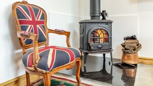 A room with a wooden chair with a Union Jack cushion, a black wood-burning stove, and a small copper pot on the floor.