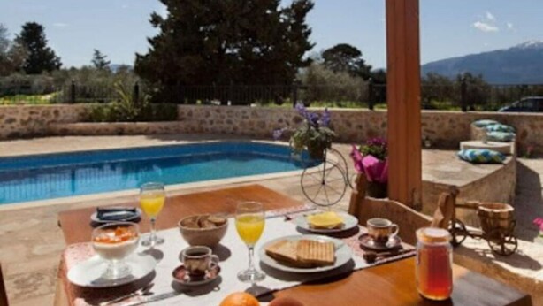 A wooden table set with breakfast items and a view of a swimming pool and mountains. - Rural Stone Villa with private pool and mountain views near local sandy beaches! (Paleloni)