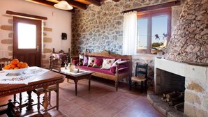 A rustic room with stone walls, a wooden ceiling, a fireplace, a sofa, a table with a bowl of fruit, and a window with curtains. - Rural Stone Villa with private pool and mountain views near local sandy beaches! (Paleloni)