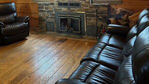 A log cabin interior with a stone fireplace, leather sofas, and a flat-screen TV mounted above the fireplace.