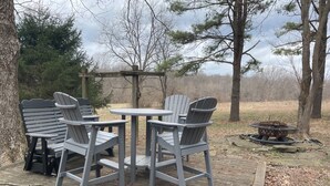 A deck with Adirondack chairs and a small round table, surrounded by trees and a fire pit.