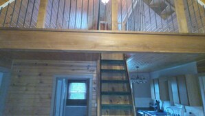 A wooden cabin interior with a loft bedroom, a kitchen area, and a staircase.