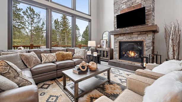 A spacious living room with a stone fireplace, leather sofa, and large windows overlooking a forest. - Trax N Trailz Lodge, Home Theatre, EV Charger, Hot Tub, Trail & Clubhouse Access (South Lawrence)