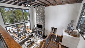 A spacious living area with a stone fireplace, large windows offering a view of trees, and a wooden ceiling structure. - Trax N Trailz Lodge, Home Theatre, EV Charger, Hot Tub, Trail & Clubhouse Access (South Lawrence)