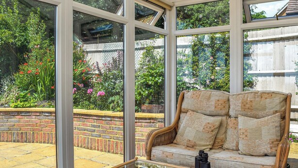 A conservatory with a wicker sofa and a glass table, surrounded by a brick wall and a garden with various plants and flowers. - A spacious and well presented bungalow with amazing views over Berkshire. (Reading)