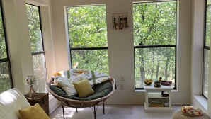 Un salon spacieux doté de grandes fenêtres, d’un ventilateur de plafond, d’un fauteuil en osier, d’un canapé blanc et d’une petite table sur laquelle se trouve un saladier rempli de fruits.