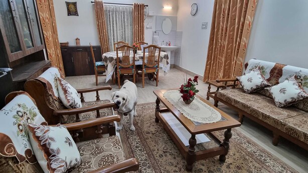 A living room with a sofa set, wooden chairs, a coffee table, and a TV mounted on the wall. - 2BR Apartment in the heart of Guwahati (Guwahati)