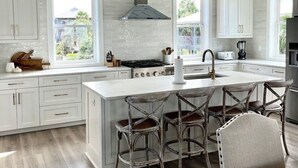 Une cuisine moderne avec des meubles blancs, un îlot central doté de tabourets de bar et une grande fenêtre offrant une vue sur l’extérieur.