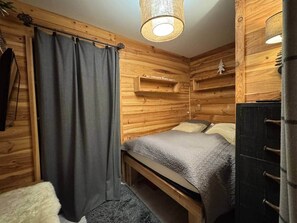 A wooden cabin-style room with a bed, a small shelf, a hanging light fixture, and a dark-colored wardrobe. - Le Tavae (Cauterets)