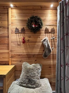A cozy room with wooden walls, a fluffy grey pillow, a grey curtain with heart patterns, a wreath with pine trees, and a red ornament.