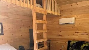 A wooden cabin room with a bunk bed, a ladder, a bed with a white sheet, a small table, and a chair.